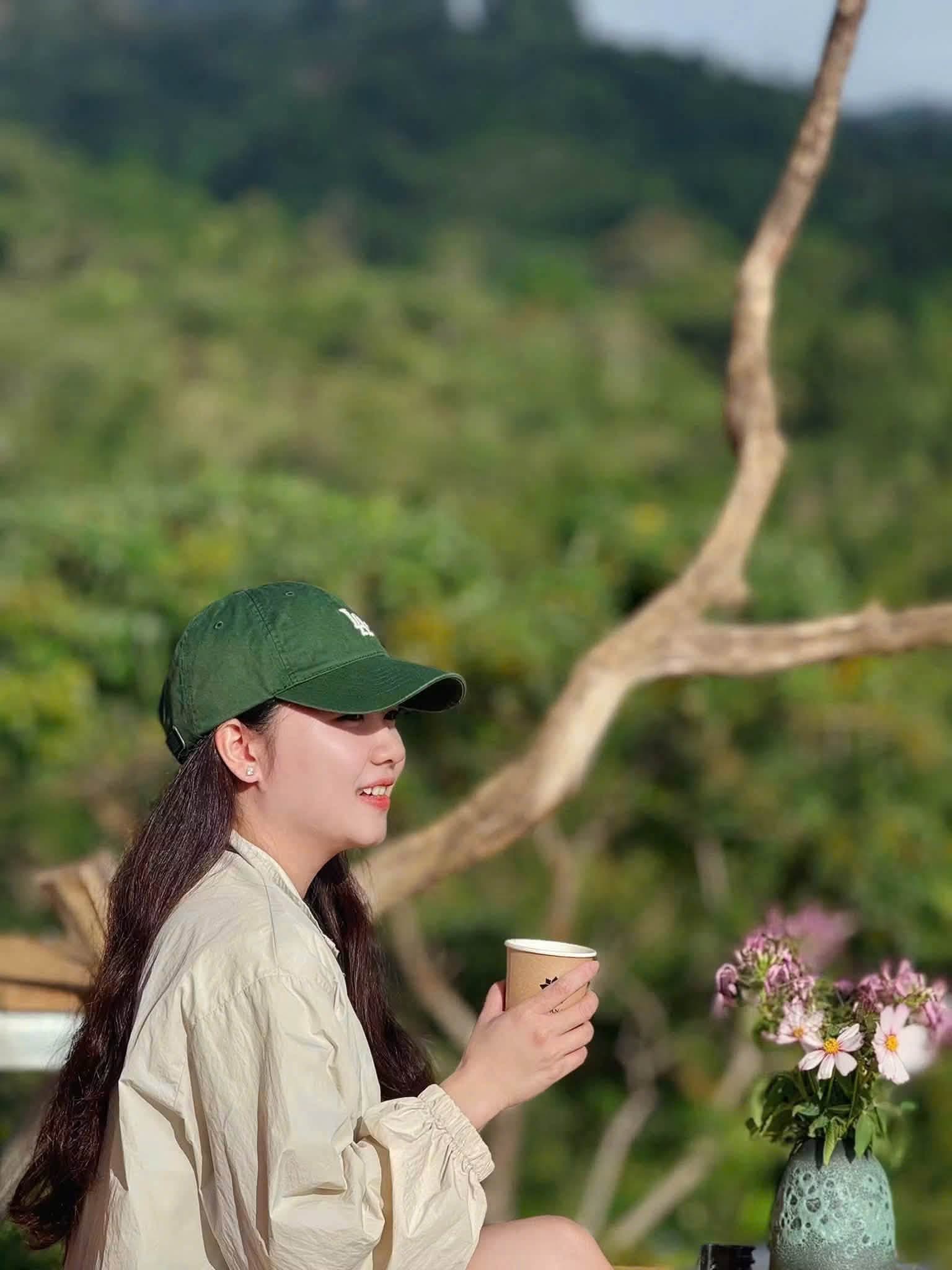 Cô gái đội mũ lưỡi trai xanh tay cầm ly cà phê giữa rừng